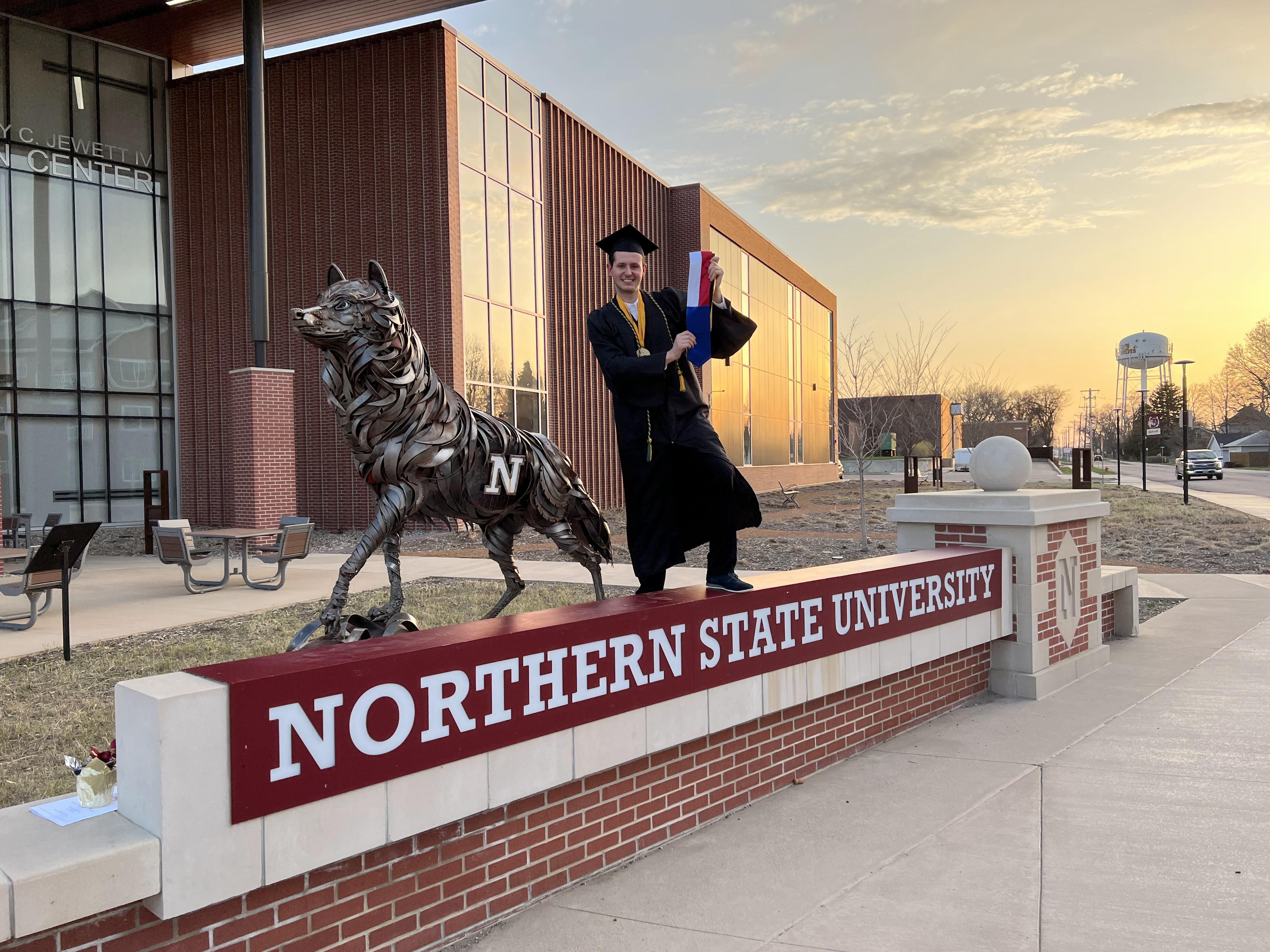 Alum Matěj Číp in cap and gown standing by the NSU Wolf statue
