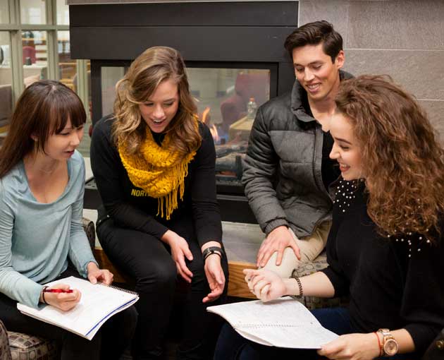 A group of smiling students looks at papers in front of a fireplace