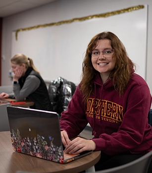 Female student working on laptop, smiling while instructor is in background