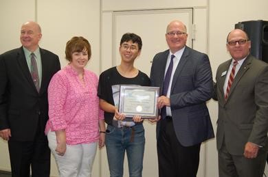 Dangju Kim Corey Klatt stands smiling with award among important BOR and NSU representatives