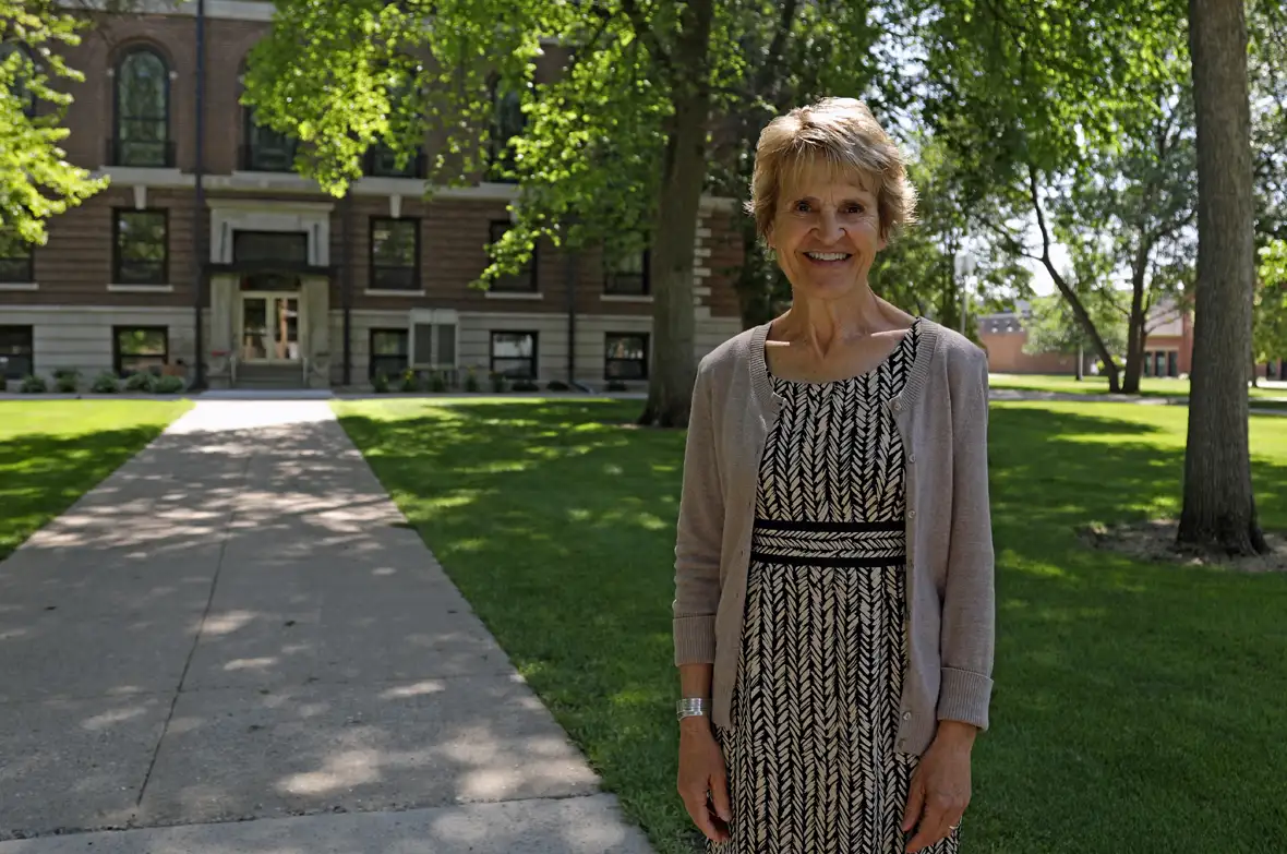 NSU Interim President Laurie Nichols, wearing a grey cardigan with a black and white patterned dress, stands on the campus green with Spafford Hall in the background.