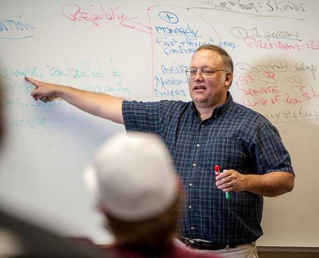 A professor in a blue shirt gestures to a whiteboard