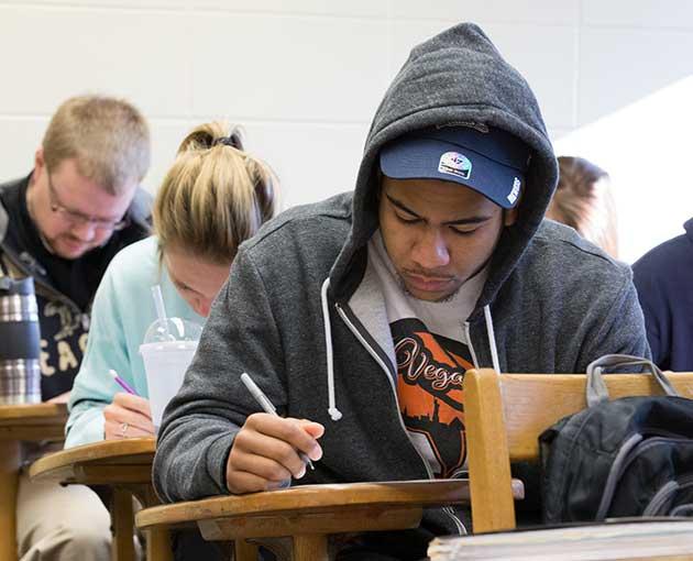 A student in cap and hoodie writes in class