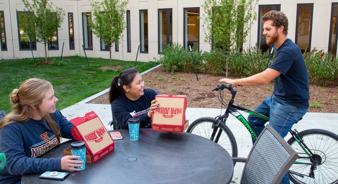 Two seated students enjoy pizza and talk to a student on bike
