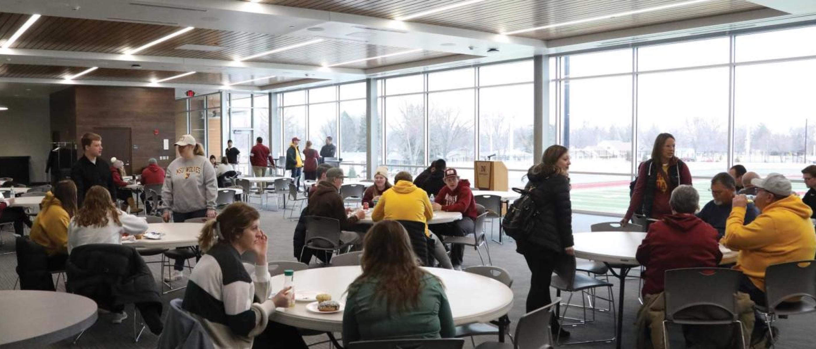 Groups of people sitting at tables in a large room with windows overlooking a football field