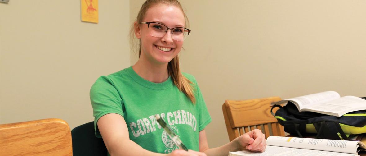 A student with glasses in a green shirt works at a table