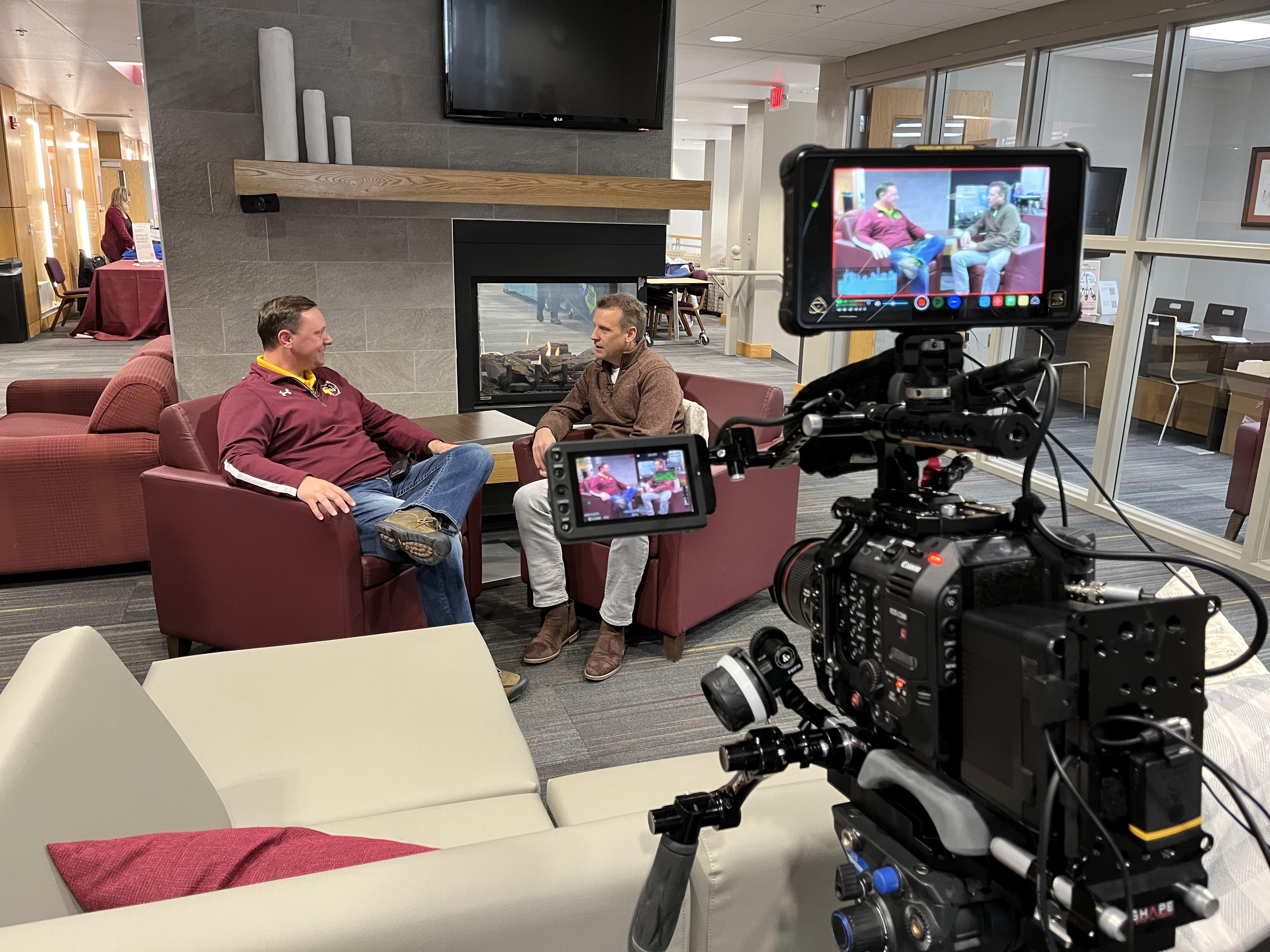Two men sitting in chairs in front of a fireplace with a camera filming their interview