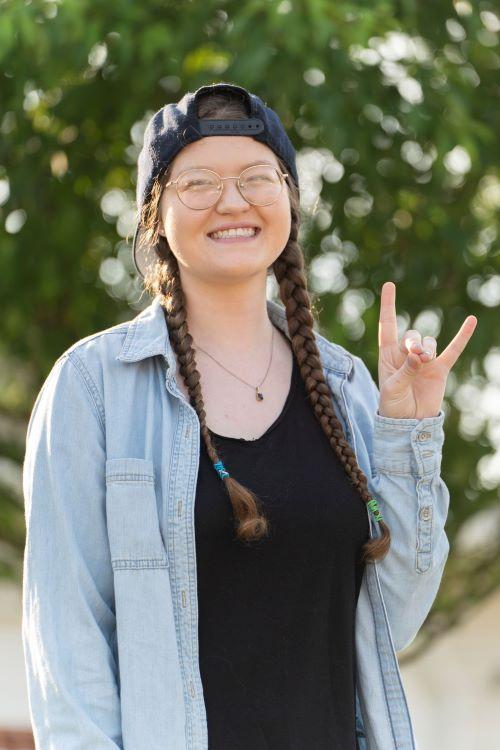 Danae Becker, in long braids, a baseball cap and denim shirt on the campus green, smiles and waves