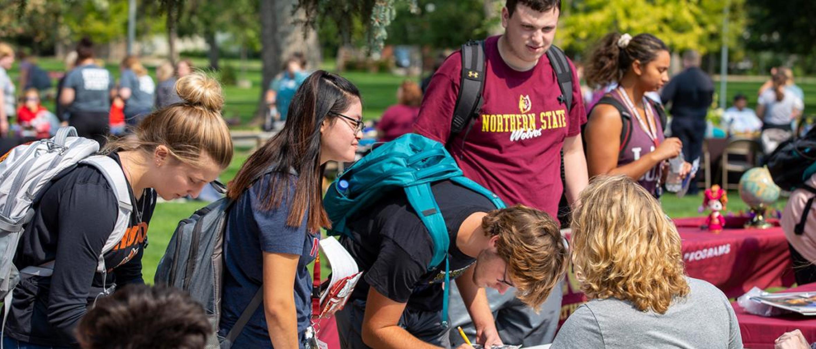 Students with backpacks surround a table on the campus green on a sunny day