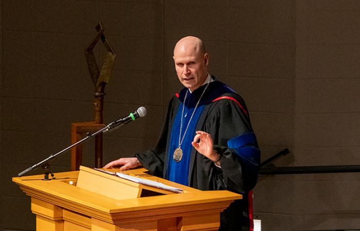 Neal Schnoor, in royal blue and black academic regalia, gestures with his left hand as he speaks at a lectern