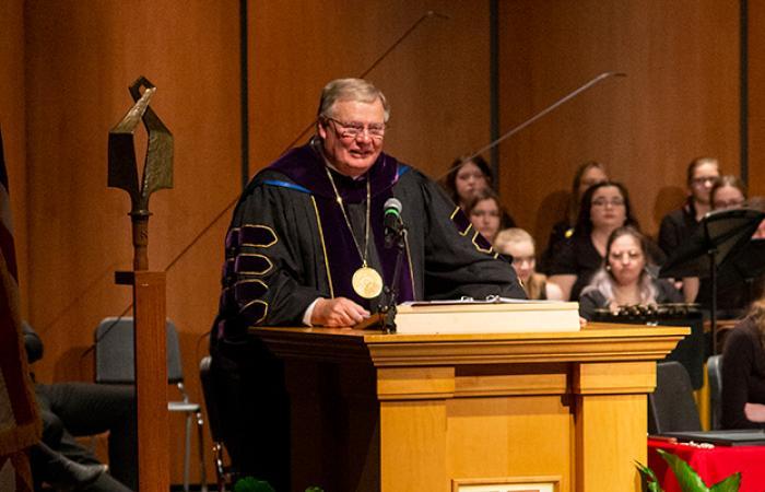 A guest in academic regalia leans on the lectern and speaks at the investiture ceremony with the NSU mace at left of photo