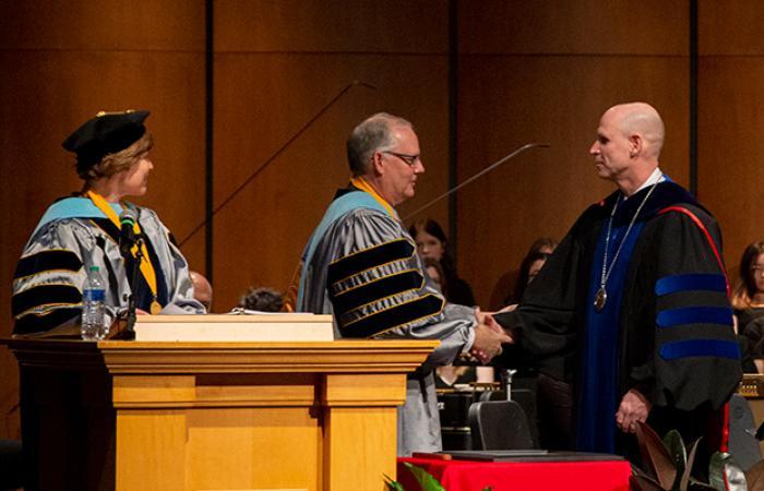 Neal Schnoor, in dark academic regalia, shakes hands with an academic guest on stage as another guest looks on
