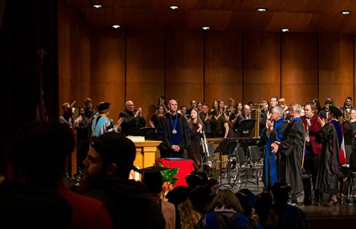 Neal Schnoor, in dark academic regalia and wearing a medallion, receives a standing ovation at the investiture ceremony