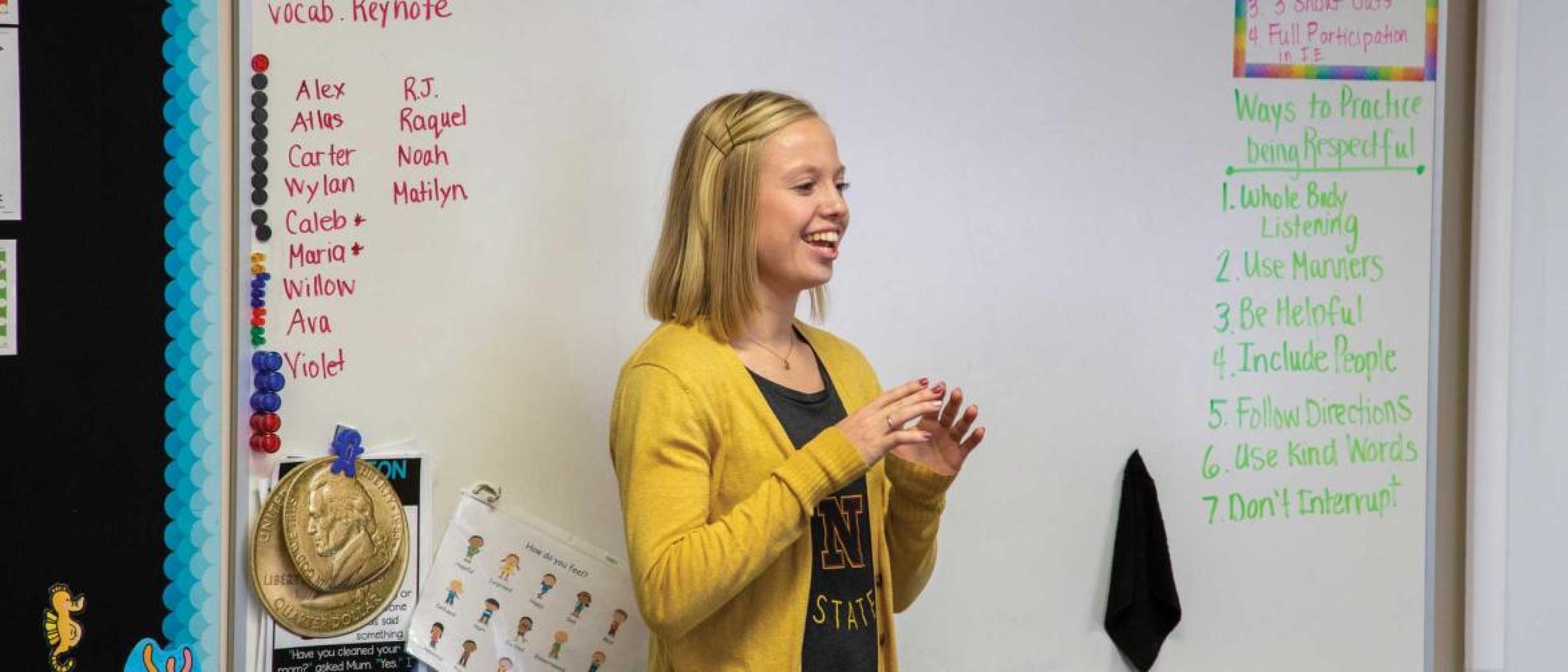  A blonde student in a yellow cardigan teaches in front of a whiteboard