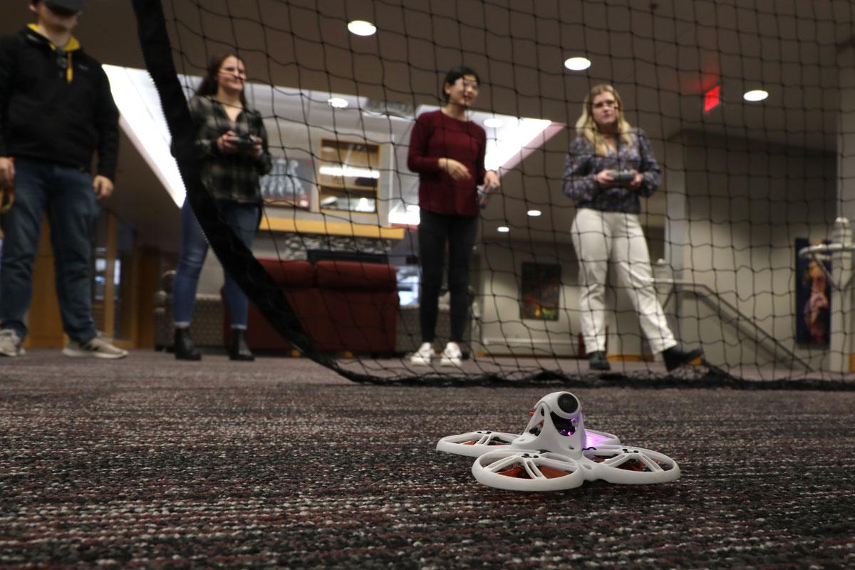 A drone rests on the floor with a net and people in the background