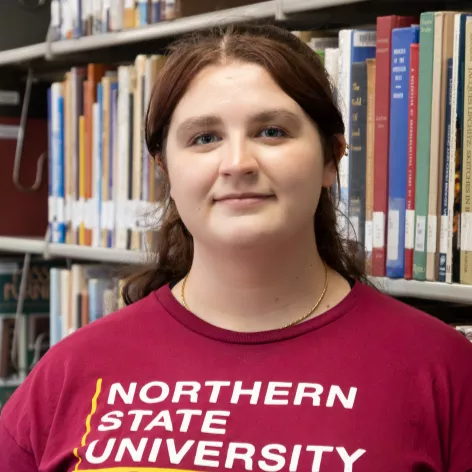 Hope Schaar standing in front of a library shelf and wearing a Northern State University shirt