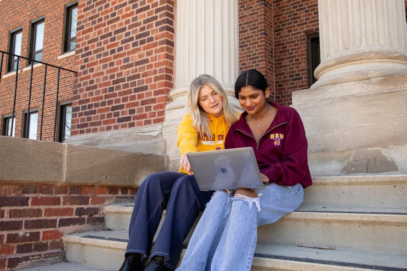 two female students sit on the steps of a building while looking at a computer