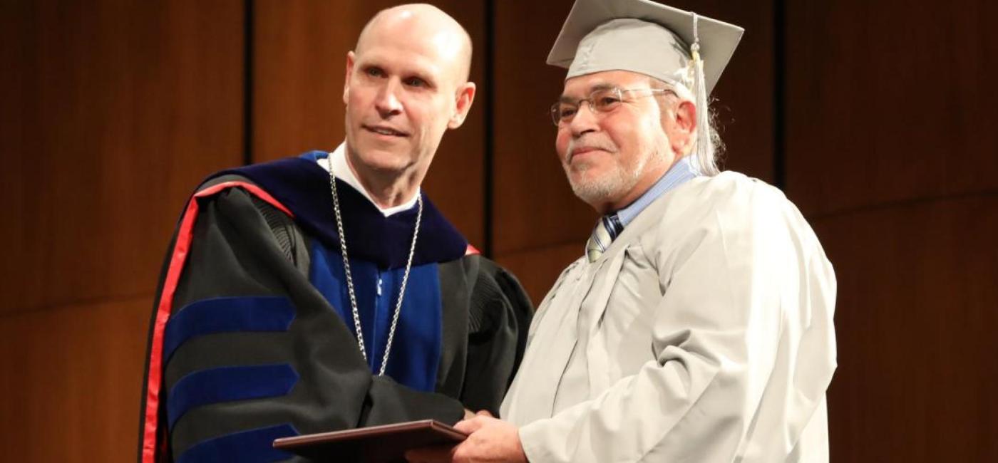 Dr. Neal Schnoor shakes hands with an older NSU graduate in a white cap and gown