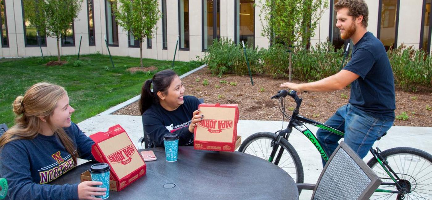 Two seated students enjoy pizza and talk to a student on bike