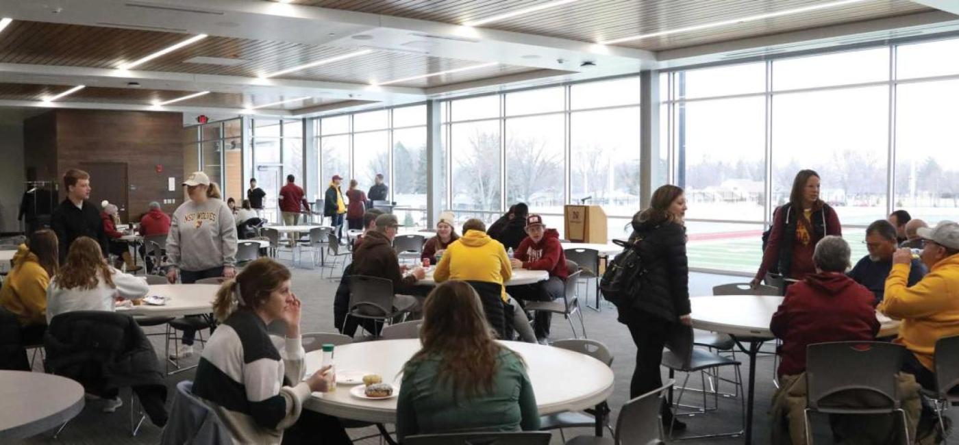 Groups of people sitting at tables in a large room with windows overlooking a football field