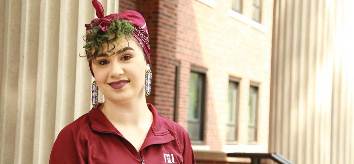 A student with green curly hair and large earrings in a Northern shirt stands outside on campus