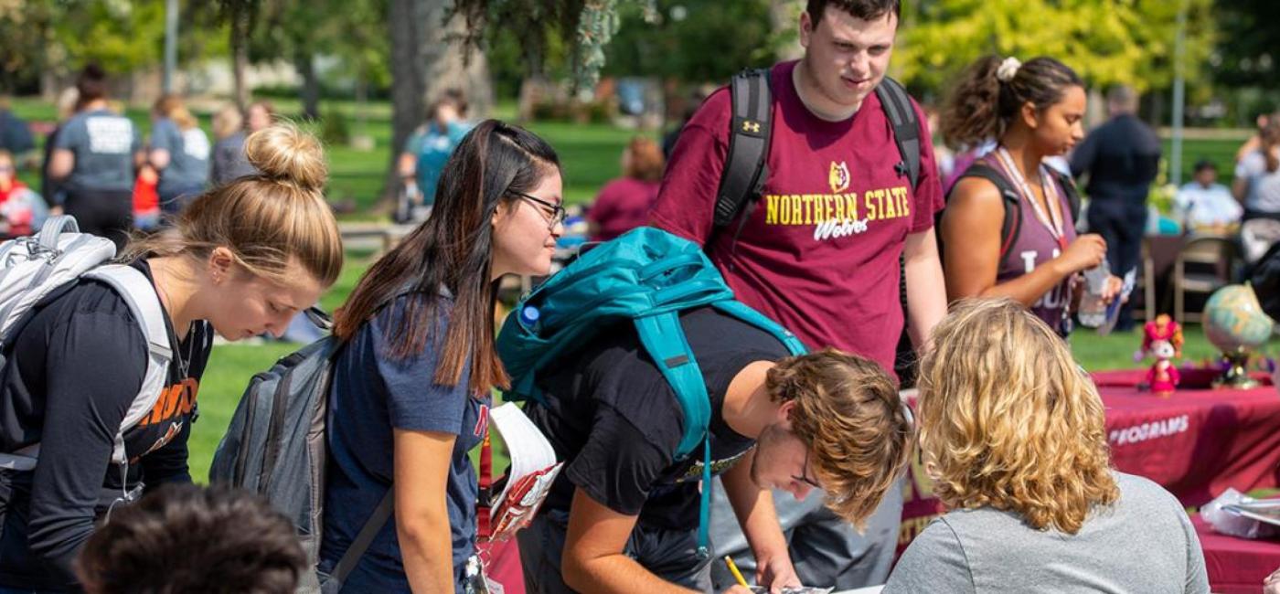 Students with backpacks surround a table on the campus green on a sunny day