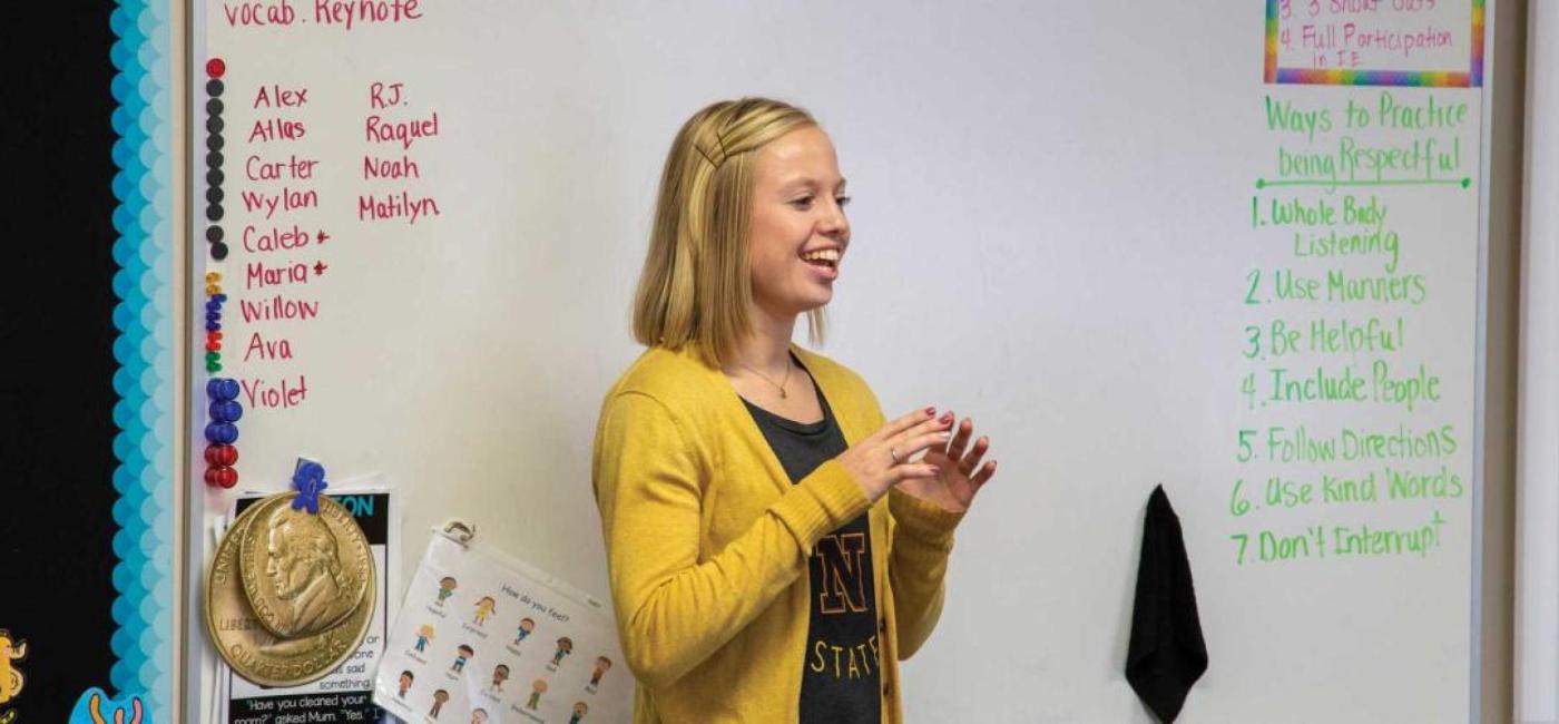  A blonde student in a yellow cardigan teaches in front of a whiteboard