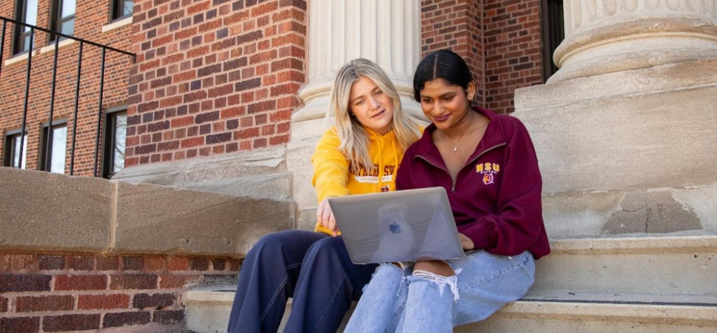 two female students sit on the steps of a building while looking at a computer