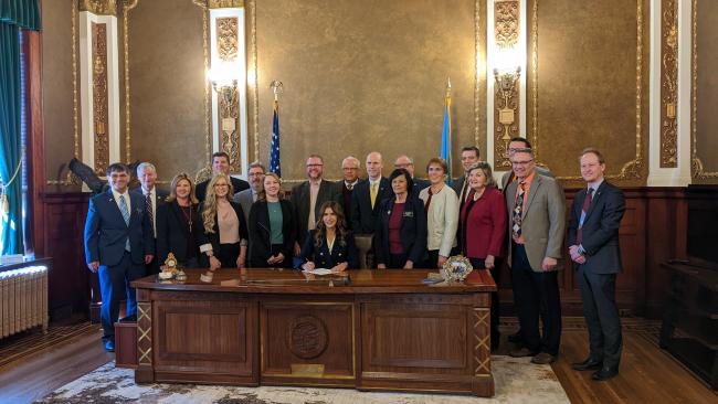 Governor Noem at her desk surrounded by local officials