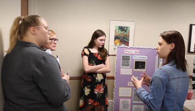 A teen girl explaining her National History Day project to two judges while another teen girl looks 
