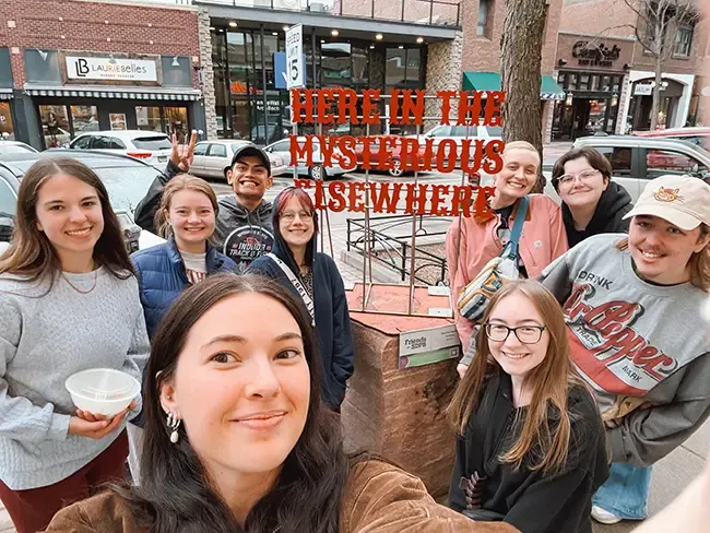 Students posing in front of a sign that says: Here in the Mysterious Elsewhere